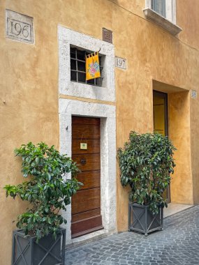 ROME, ITALY - 30 Temmuz 2025 Narrow sokak manzarası üzerinde ahşap kapılı ve renkli bir bayrak asılı eski sarı bir bina cephesini gösteriyor. Girişi çevreleyen iki saksı yeşili.