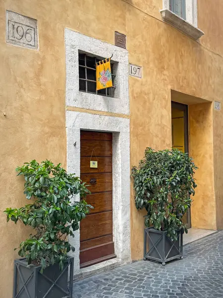 ROME, ITALY - 30 Temmuz 2025 Narrow sokak manzarası üzerinde ahşap kapılı ve renkli bir bayrak asılı eski sarı bir bina cephesini gösteriyor. Girişi çevreleyen iki saksı yeşili.
