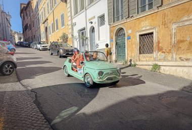 ROME, ITALY - 1 AĞUSTOS 2025 Cobblestone yolunda bir grup mutlu turist taşıyan canlı bir turkuaz klasik araba yokuş aşağı gidiyor. Sokaklar tarihi binalarla dolu.