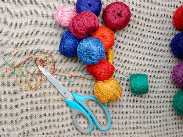 unwound threads from multi-colored skeins and scissors on gray burlap. multi-colored balls of thread and scissors
