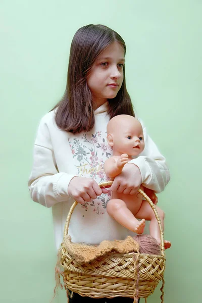 a female child holds a doll and a wicker basket with yarn in her hands. Knitting yarn and a doll in the hands of a female child