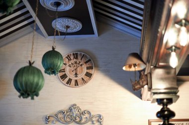 Fotoğraf, içinde retro estetik olan bir iç mekan gösteriyor. Odak noktası, üzerinde Roma rakamları olan tahta bir duvar saati. 