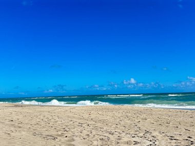 Yeşil renkli okyanus dalgalarının yavaşça kıyıya vurduğu kumlu bir sahil manzarası. Gökyüzü açık deniz manzarasına derinlik katan dağınık beyaz bulutlarla parlak mavi.. 