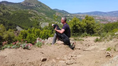 Bir adam bir köpeğe sarılır ve ona dağların güzelliğini ve doğayı gösterir..