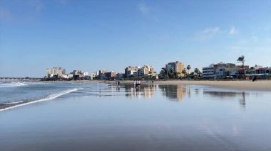 Peru 'daki Pimentel sahili sabah saatlerinde rastgele plaj yaşamını gösteriyor. İnsanlar sahilde yürüyor, sabah melteminin tadını çıkarıyorlar. Arka planda evler ve rıhtım var. Okyanus, turist mekanının güzel ayna efektini yapıyor.