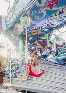 Şehir panayırının önünde oturan renkli kıyafetli güzel bir genç kadın eğlence parkının hünerlerinin tadını çıkarıyor..