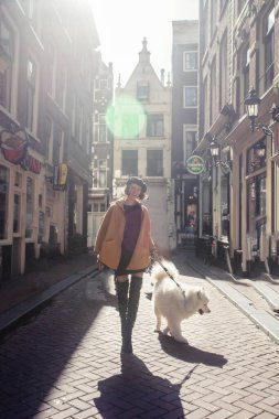 Amsterdam sokaklarında beyaz köpekle yürüyen güzel bir kadının portresi. Model yüksek botlar, zarif bir ceket ve bere giyiyor. Doğal güneş ışığı etkisi.