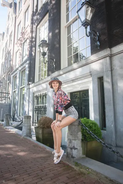 Amsterdam 'ın şık sokaklarında poz veren güzel, mutlu genç bir kadın. Moda modeli neşeli, mutlu ve şehir seyahatinden keyif alıyor. Tahta şapka, yamalı bomba ceketi, şort ve spor ayakkabı giyiyor..