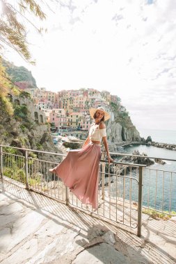 Modayı takip eden genç bir kadın çitte mutlu bir şekilde oturuyor. Gülümsüyor ve İtalya 'nın Cinque Terre Manarola şehrinde turistik manzaranın tadını çıkarıyor. Uzun pembe ipek etek ve şapka giyiyor..