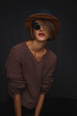 Siyah arka planda, şapka, güneş gözlüğü, kırmızı dudak ve süveter giyen manken portresi. Saçları yarı yarıya örtülü ve kameraya bakan güzel bir kadının vesikalığı..