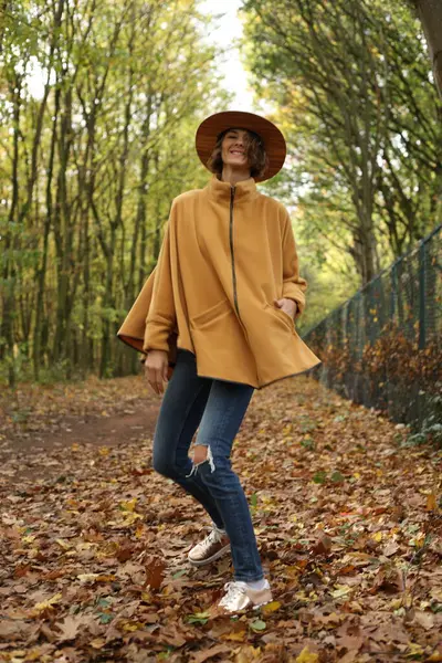 Şapka takan, şık bir panço ceket giyen güzel bir kadın yürüyor ve sonbahar ormanı manzaralı bir yerde eğleniyor. Sonbahar mevsimi konsepti