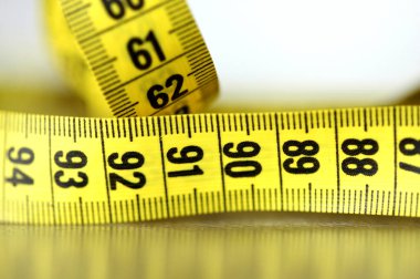 Beyaz arka planda sarı bir ölçüm bandına yakın plan, altında yansıma var. Mükemmel ölçüler 60-90-60 sembolik fitness, model gerekliliği, vücut idolü, dikiş, diyet, sağlık, beden bilinci, ama kadın için de baskı.