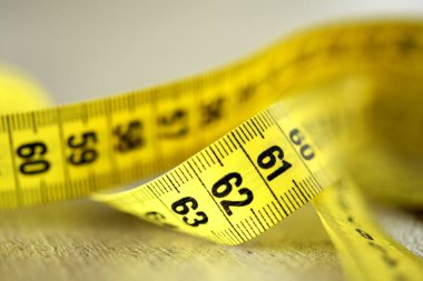 Beyaz arka planda sarı bir ölçüm bandına yakın plan, altında yansıma var. Gerçekçi olmayan bel ölçüsü, sembolik fitness, model gerekliliği, vücut idolü, dikiş, diyet, sağlık, beden bilinci, ama kadın için de baskı.