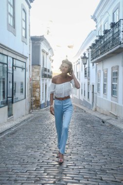 Portekiz, Faro sokaklarında, saçlarına dokunurken kameraya doğru yürüyen güzel bir genç kadın. Şık bir model, şık bir yaz elbisesi, omuzları açık ve çizgili elbiseler giyiyor. Moda seyahat konsepti.