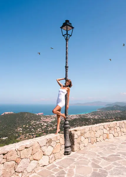 Korsika 'da Akdeniz manzarası önünde beyaz kısa elbiseli, eski moda sokak lambasına poz veren güzel bir kadın. Avrupa 'da yaz aylarında seyahat eden moda modeli.