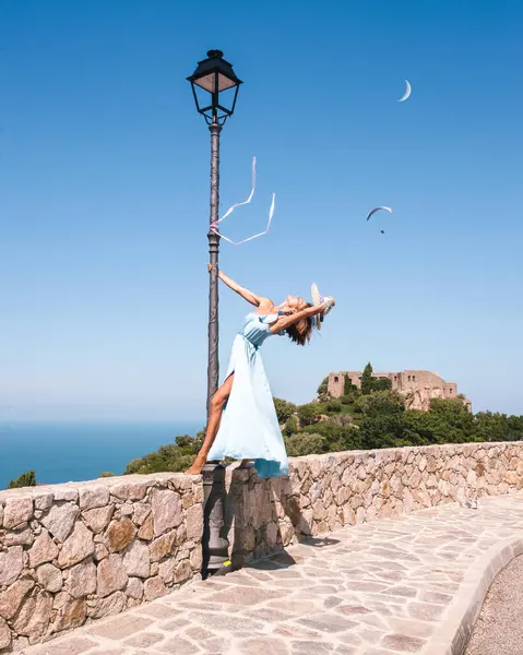 Duvarda, eski moda sokak lambasının yanında duran güzel bir kadın Korsika 'da Akdeniz' in önünde uzun mavi bir elbiseyle dans ederken arka planda küçük bir şato.. 