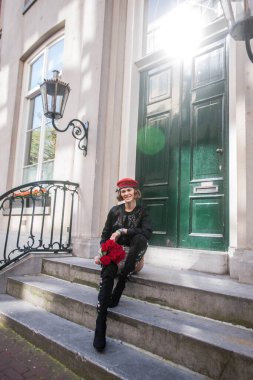 Amsterdam sokaklarında zarif yeşil kapıların önünde merdivenlerde duran şık bir kadın. Moda mankeni elinde güllerle kameraya gülümsüyor. Moda seyahati kavramı