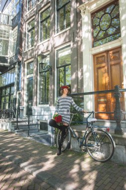 Güzel, zarif bir kadın Avrupa 'nın tarihi Amsterdam şehrinde yaz tatilinin tadını çıkarırken siyah beyaz örgü elbiseler, kırmızı askeri şapkalar ve bisiklet önünde poz veren kırmızı güller giyiyor..