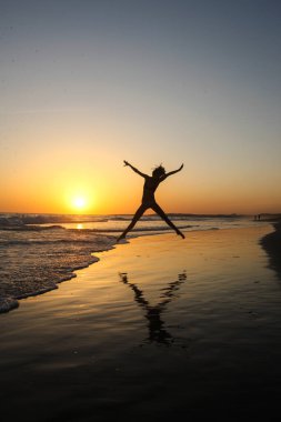 Portekiz 'de denizin üzerinde güzel altın gün batımının önünde kolları ve bacakları ayrık bir şekilde muhteşem bir atlayış yapan ince kadın silueti. Aktif yaşam tarzı pozitif duygular, gün doğumunu ve günbatımını kovalamak