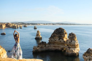 Uzun yaz çiçekli elbisesiyle tanınmayan bir kadın kollarını kaldırmış ve Portekiz Lagos 'taki güzel kaya oluşumlarını ve denizleri seyrediyor..