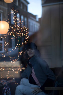 Işıklı Noel ağacının yanında elinde bir dergi tutarken gülümseyen güzel bir kadının dikey fotoğrafı. Zarif Noel atmosferiyle çevrili kendine güvenen şık bir kadının penceresinden güzel bir portre..