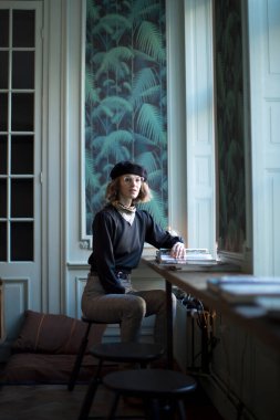 Genç ve güzel bir kadın pencerenin önündeki masada oturmuş kameraya ciddi ciddi bakarken kitap okuyor. Edebiyat, eğitim, zeka, moda, dergi, menü, bilgi, doğal aydınlatma