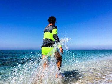 Neon spor giyimli sporcu Sardinya 'da turkuaz renkli suyun yanındaki plaja koşuyor. Harekete geçen sporcu, koşarken etrafa su sıçratıyor.