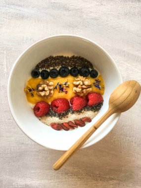 Renkli meyve, tohum ve tahta kaşıkla süslenmiş sağlıklı bir kahvaltı kasesi. Yunan yoğurdu, chia, yabanmersini, ceviz, mango püresi, ahududu, susam ve goji 'den yapılmış sabah yemeği için nefis bir fikir..