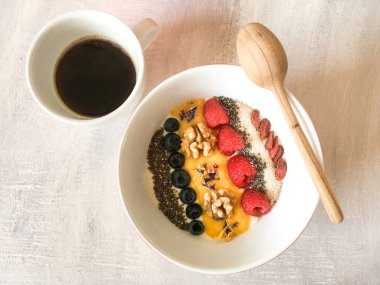 Renkli meyve, tohum ve ahşap kaşıkla süslenmiş kâsenin yanında sade sağlıklı kahvaltı kahvesi. Yunan yoğurdu, chia, yaban mersini, ceviz, mango püresi, ahududu, susam...