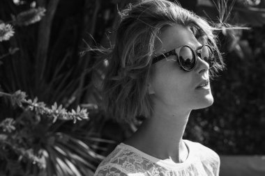 Beyaz dantelli, güneş gözlüklü zarif bir kadının yatay tek renkli portresi. Siyah ve beyaz. Moda modelinin doğal güneş ışığıyla çekilmiş yakın plan fotoğrafı.. 