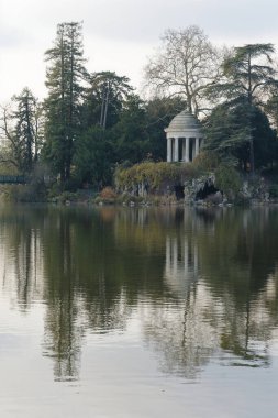 Daumesnil Gölü 'nün dingin sularında kusursuz bir şekilde yansıtılan ve Bois de Vincennes' in huzurlu atmosferini gösteren kubbeyi ve yapay mağarayı sakin bir şekilde yakalar..