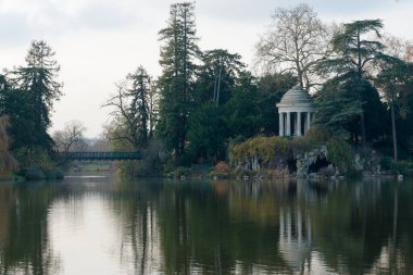 Bois de Vincennes 'deki Daumesnil Gölü' nün yanında bulunan ve yemyeşil ağaçlar ve sakin sularla çevrili olan kubbe ve yapay mağarayı görmektedir..