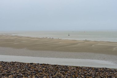 Geniş bir kumlu sahil, bulutlu gökyüzünün altında alçak gelgitte ıslak bölgeler ortaya çıkarır. Uzak tekneler sakin sularda, sakin bir kıyı manzarası tarafından çerçevelenmiş, ihtiyatlı bir şekilde yol alırlar..