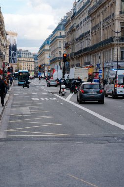 Paris, Fransa-2024-11-20 yoğun bir cadde boyunca trafik akışı araba, otobüs ve scooterlarla harmanlanır. Yayalar bulutlu gökyüzünün altındaki tarihi binaların yanında yürürler.