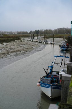 Birkaç tekne bir nehir kıyısına demirlemiş ve sular çekildiği için çamurlu kıyılar açığa çıkmış. Sakin su bulutlu gökyüzünü yansıtıyor, kırsal bölgede sakin bir atmosfer yaratıyor..