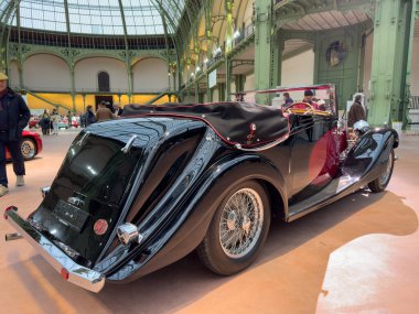 Paris,France-04.02.2025 A vintage convertible glistens under the bright lights of an exhibition space, showcasing its timeless design and gleaming chrome details amid a stylish gathering. clipart