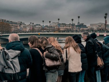 Bir grup insan Paris 'te nehir kıyısında sıcak kıyafetler giyip serin ve bulutlu bir akşamın tadını çıkarıyorlar. Suyun karşısındaki şehre bakıyorlar, canlı bir atmosfer yaratıyorlar..
