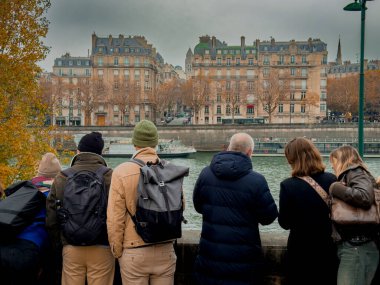 Turist grupları, serin bir sonbahar gününde tarihi mimariye hayran kalarak Seine Nehri boyunca toplandılar. Altın ağaçlar Paris 'in ikonik ufuk çizgisinin manzarasını çerçeveliyor.