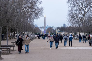 Ziyaretçiler, soğuk havanın ve uzaktaki uzun dikilitaşın manzarasının tadını çıkararak çıplak ağaçlarla kaplı geniş bir patika boyunca yürürler. Park hareketlilikle dolup taşıyor.