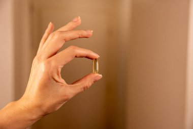 Bir el, yumuşak ve nötr bir arkaplana karşı altın bir omega 3 kapsülü dikkatlice tutuyor. Işık kapsüllerin parıltısını vurguluyor, sağlık yararlarını ve sağlığını temsil ediyor. Bu an şunu vurguluyor: