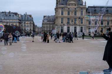 Bir sanatçı serin havanın tadını çıkaran ziyaretçilerle dolu kalabalık bir parkta büyük ve küçük kabarcıklar yaratır. Zarif mimari, eğlenceli aktiviteler için çarpıcı bir zemin sağlar..