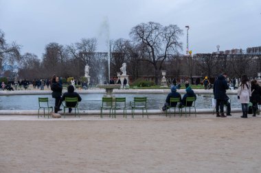Ziyaretçiler, bulutlu bir gökyüzüne karşı çıplak duran ağaçlar sakin bir atmosfer yaratırken, şehir parkındaki bir çeşmenin yanında huzurlu bir anın tadını çıkarıyorlar..