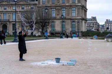 Koyu renk giyinmiş bir insan, serin ve bulutlu bir günde, tarihi mimariyle çevrili bir bahçede büyük baloncuklar yaratıyor. Vazolar ve kovalar yakında..