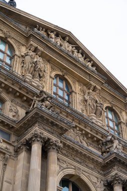 Louvre Müzesi 'nin çarpıcı mimari özellikleri ayrıntılı heykelleri ve dekoratif unsurları ön plana çıkarıyor. Ziyaretçiler açık bir gökyüzü altında sanatsal işçiliğe hayret ediyorlar.