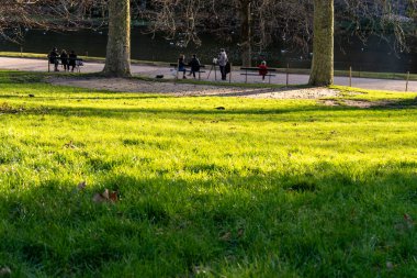 İnsanlar Parc des Buttes-Chaumont 'ta çimlerin üzerinde güneşli bir öğleden sonranın tadını çıkarıyorlar. Bazıları sohbete dalarken, diğerleri su kenarındaki manzaranın tadını çıkarıyor..