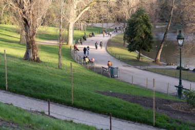 İnsanlar Paris 'teki Parc des Buttes-Chaumont' un kıvrımlı yolları boyunca sakin bir şekilde yürürler. Parkta yuvarlanan yeşil çimenler, ağaçlar ve sakin bir atmosfer yer alıyor..