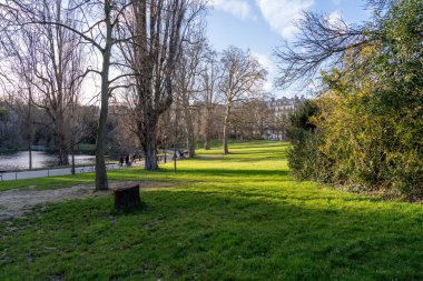 Ziyaretçiler, güneşli bir günde Parc des Buttes-Chaumont 'un yemyeşil alanlarında dolaşır ve canlı manzaranın ve huzurun tadını çıkarırlar. Parkta ağaçlar ve huzurlu bir göl sergileniyor..