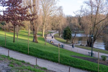 Ziyaretçiler, Parc des Buttes-Chaumont 'ta, ağaçlar ve dingin bir gölet ile çevrili dolambaçlı yollarda yürürler. Parktaki güzel bir bahar gününün özünü yakalarlar..