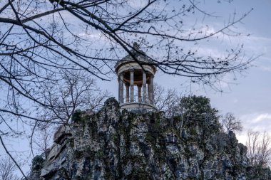 Belvedere yapısı Parc des Buttes-Chaumont 'taki kayalık bir tepenin tepesinde alacakaranlık yaklaşırken, dallar manzarayı çerçeveliyor ve sakin bir atmosfer yaratıyor..