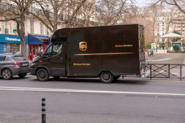 Şehirde hareketli bir şehir caddesi boyunca büyük bir teslimat kamyonu park halinde. Koyu renk şemasını içeren kamyon, park halindeki bir sıra arabanın yanında yer alıyor. Aracın etrafında yayalar var.